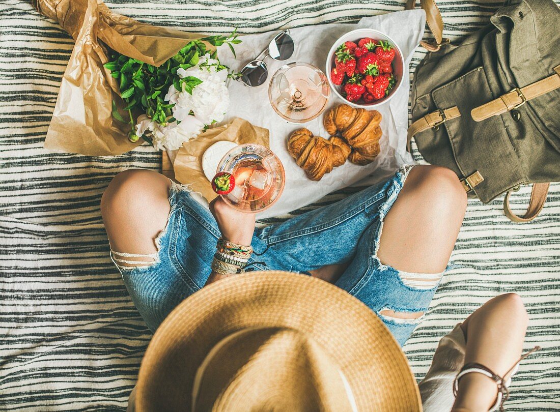 A woman wearing ripped jeans holding a glass of wine next to strawberries and croissants on a picnic blanket (seen from above)