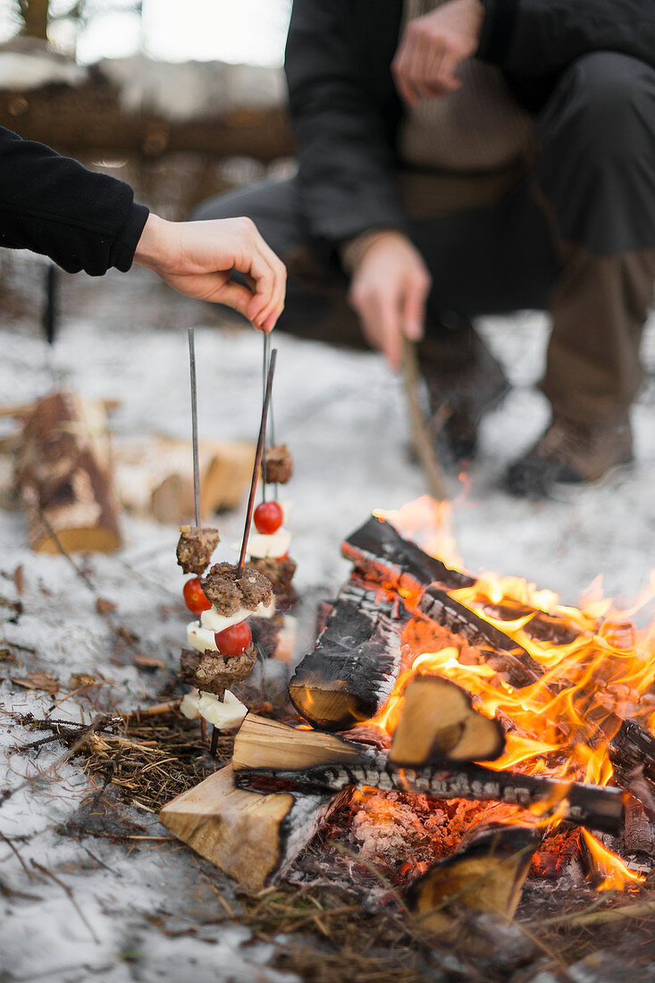 Two people cooking skewers by campfire