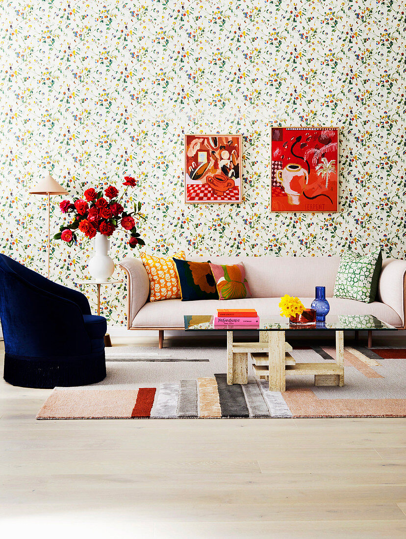 Upholstered sofa, side table with bouquet of roses, armchair and coffee table in the living room with floral wallpaper, pictures on the wall