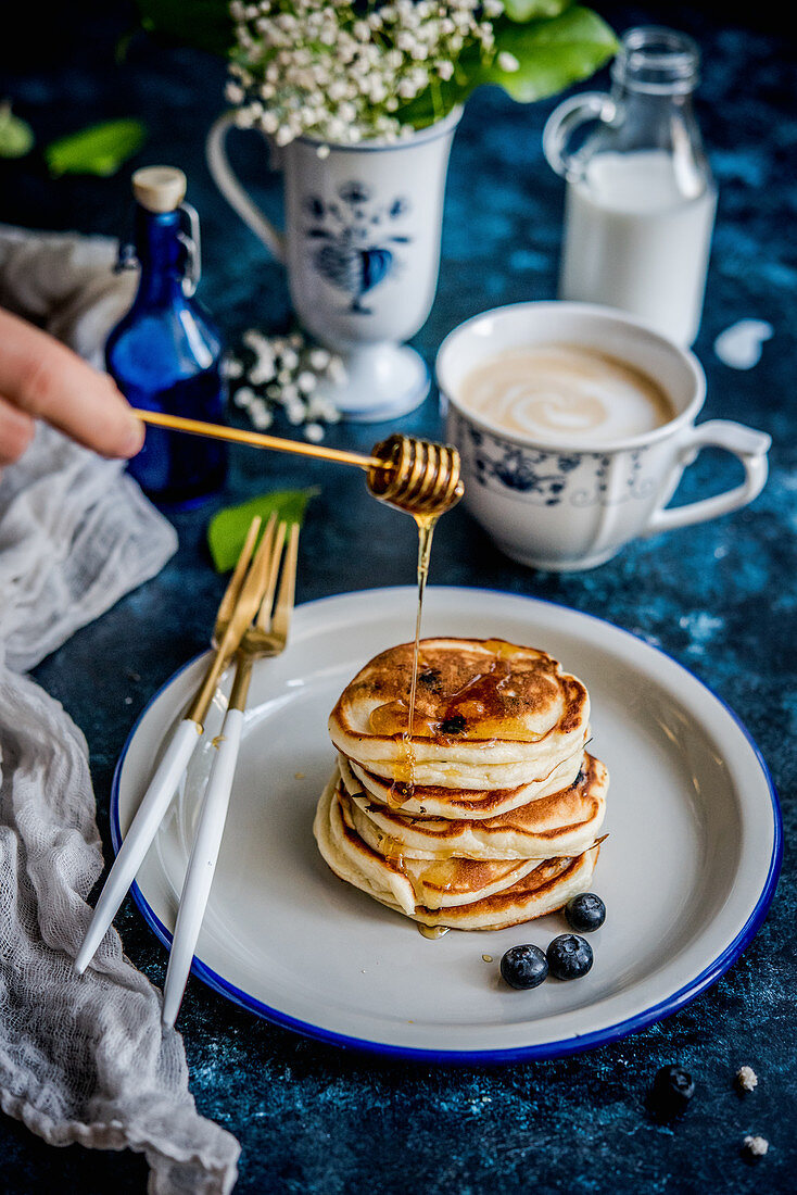 Gestapelte Blaubeer-Pancakes mit Honig vor einer Tasse Milchkaffee