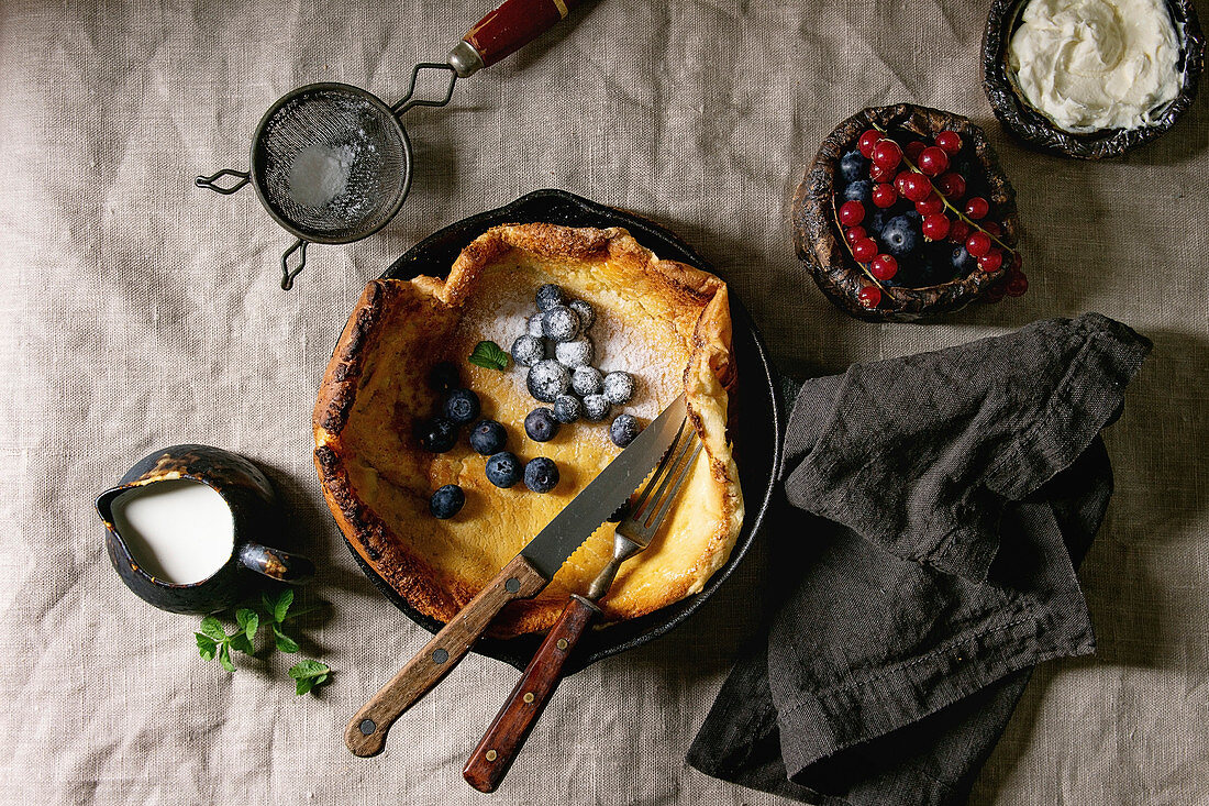 Fresh baked Dutch baby pancake in iron cast pan served with blackberry and red currant berries, mascarpone cheese, sugar powder, jug of cream