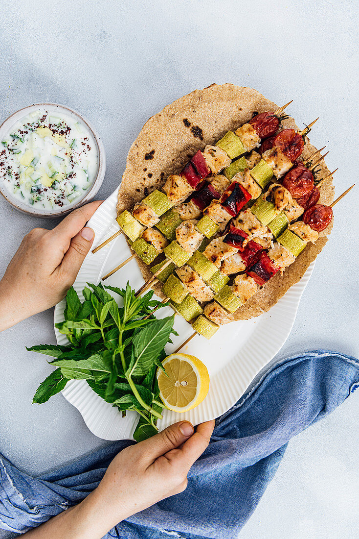 Woman serving spicy chicken kabobs on lavash bread accompanied by herbs, yogurt dip and lemon