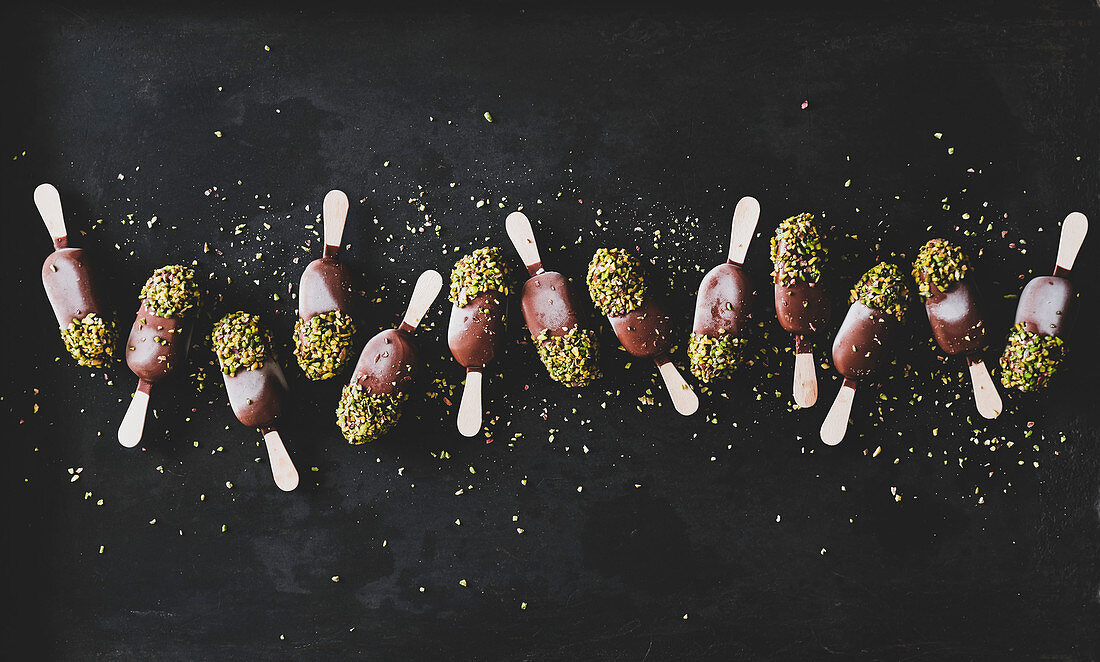 Ice cream popsicle pattern: Flat-lay of chocolate glazed ice cream pops with pistachio icing over dark background