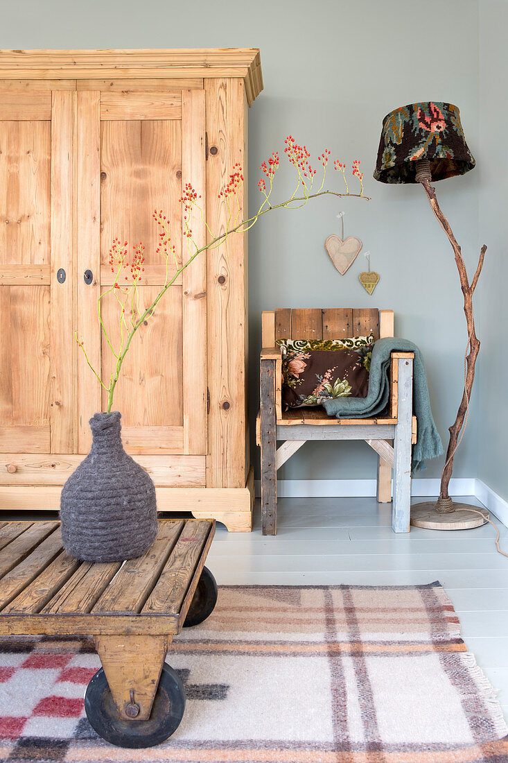 Branch of berries in demijohn with felted cover on rustic wooden table, wardrobe, wooden chair and standard lamp in living room