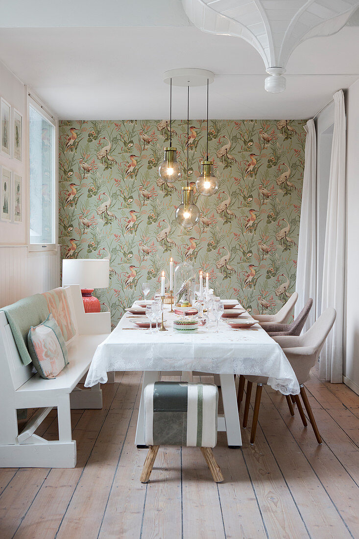 Festively set table, white bench and chairs in dining room with bird-patterned wallpaper
