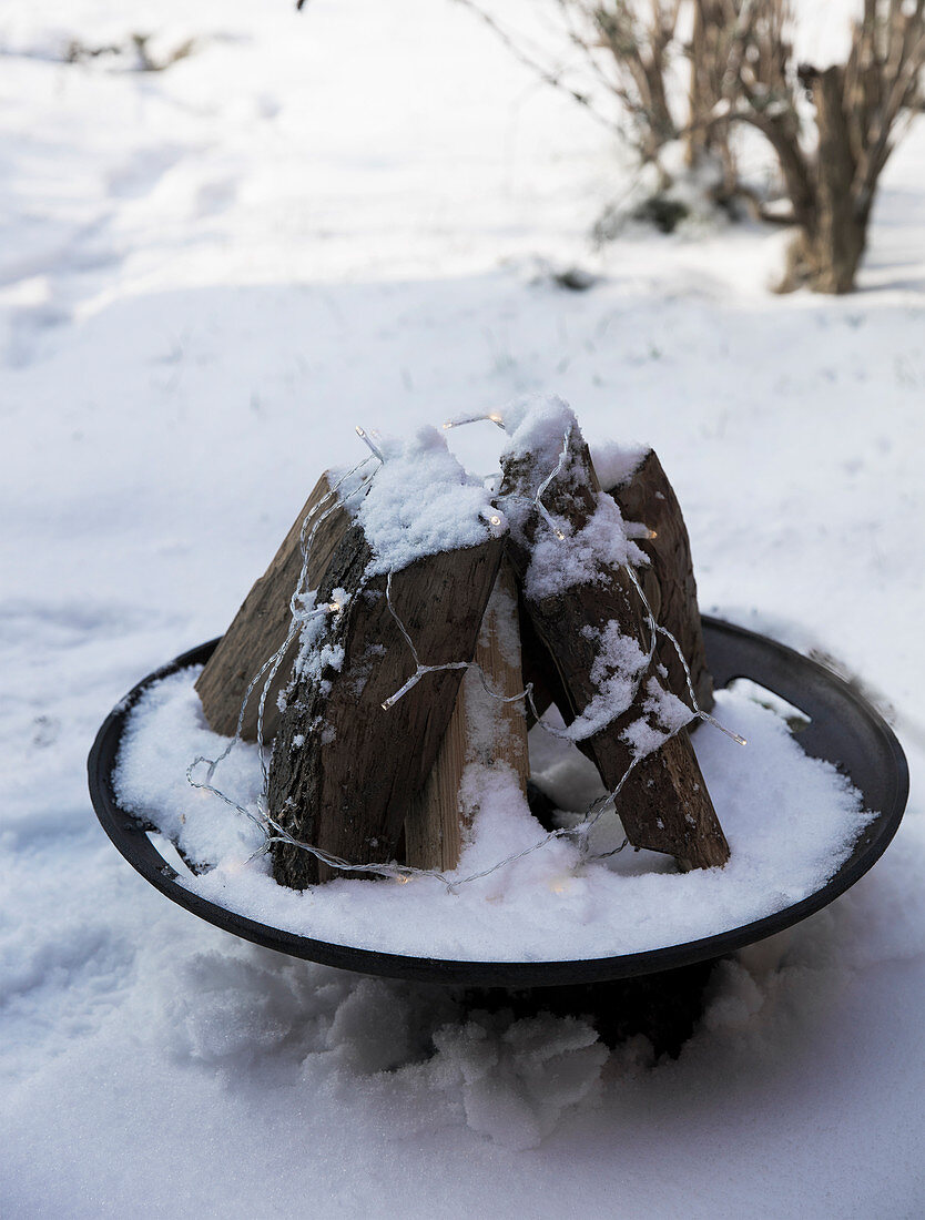 Firewood in a fire bowl with fairy lights in a snowy garden