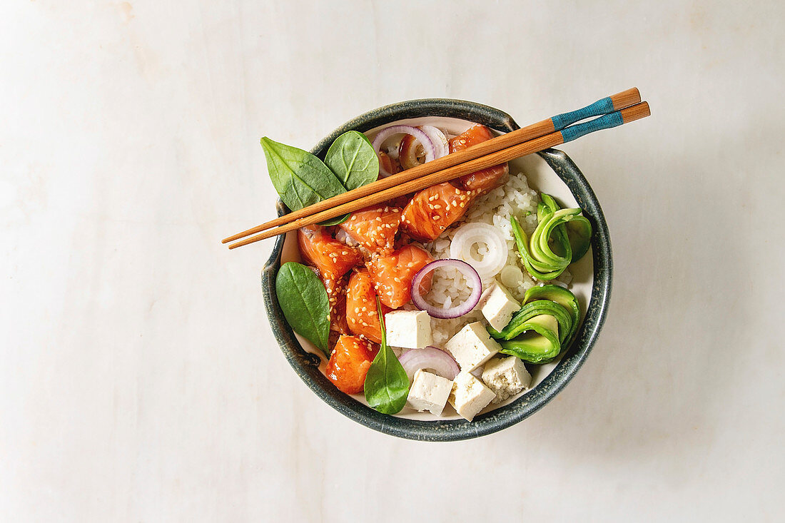 Poke bowl with soy sauce marinated salmon, rice, avocado and tofu cheese served in ceramic bowl with chopsticks