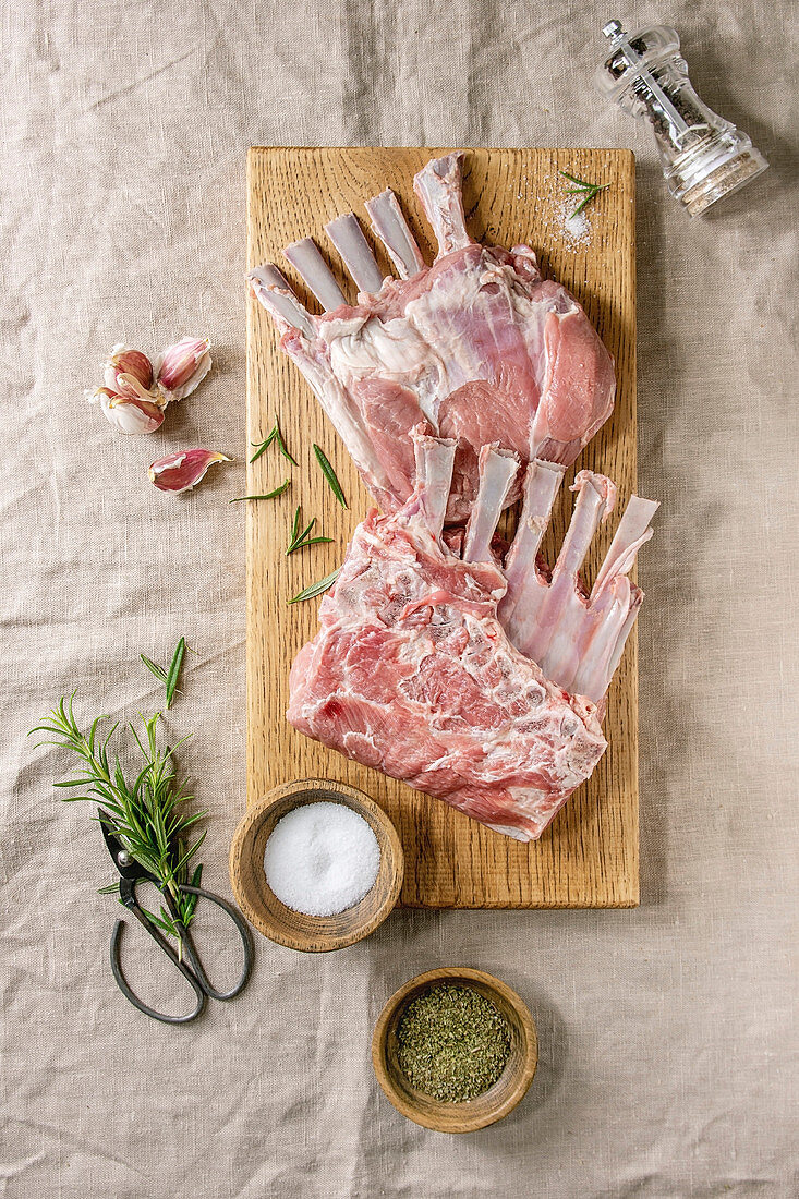 Raw uncooked rack of lamb on wooden cutting board with salt, herbs rosemary, pepper and garlic