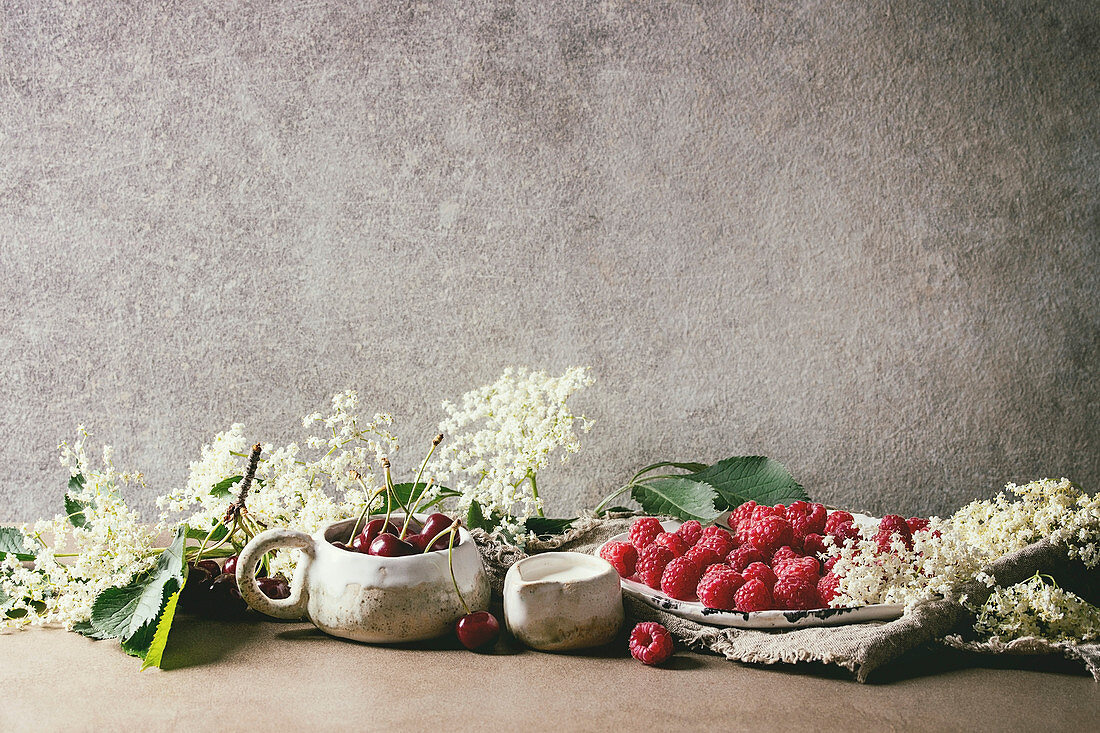 Fresh cherry and raspberry berries in ceramic mug and plate, elderflowers, jug of cream on kitchen table