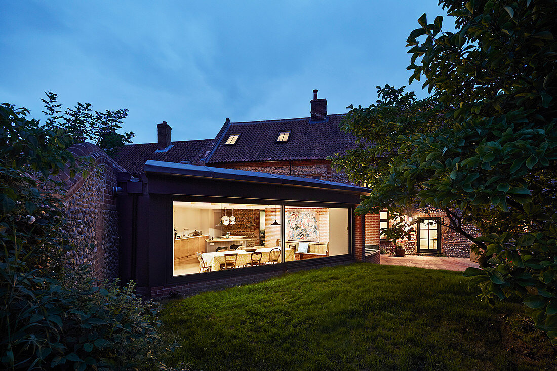 Blick vom Garten durch Glasfront in beleuchteten Anbau mit Essbereich bei Abenddämmerung