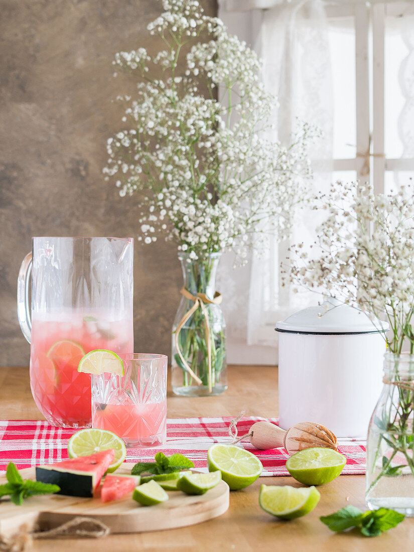 Cold glass jug of iced pink lemonade with watermelon and lime slices on rustic kitchen table and bunch of gypsophila flowers