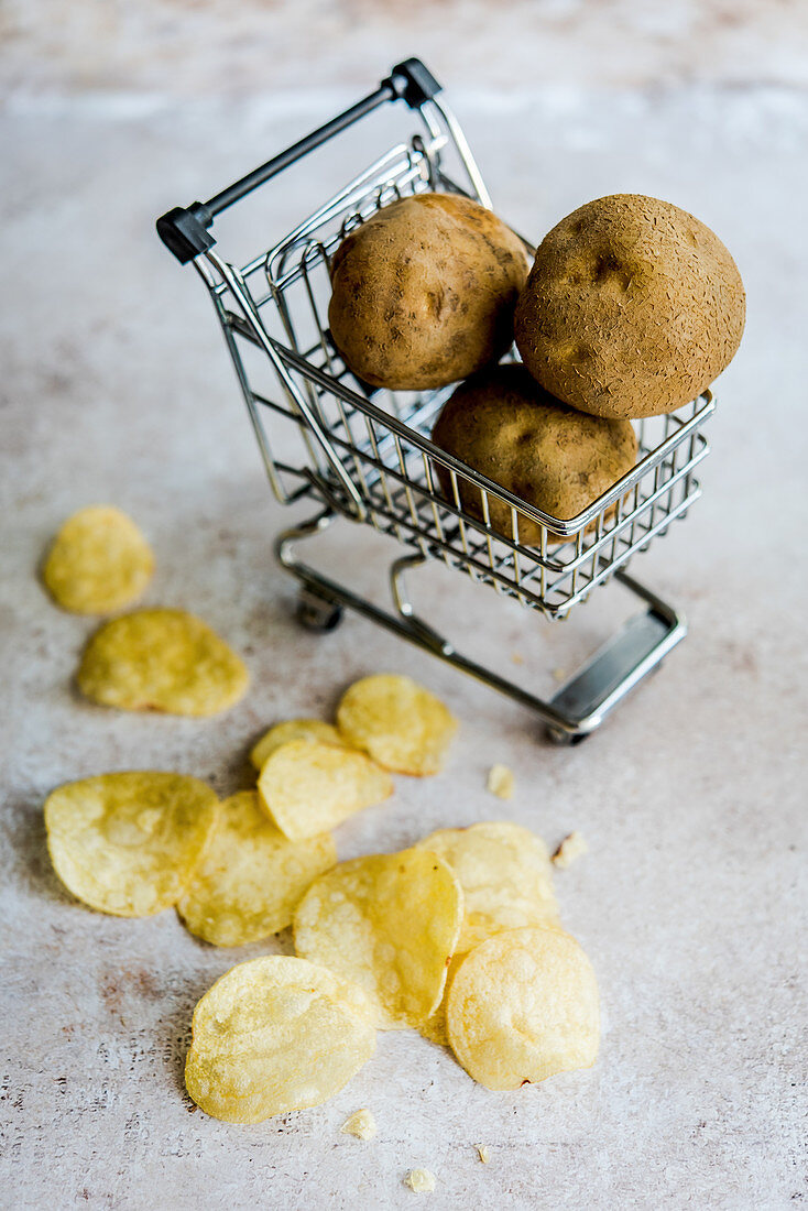 Kartoffelchips, dahinter rohe Kartoffeln in Miniatur-Einkaufswagen