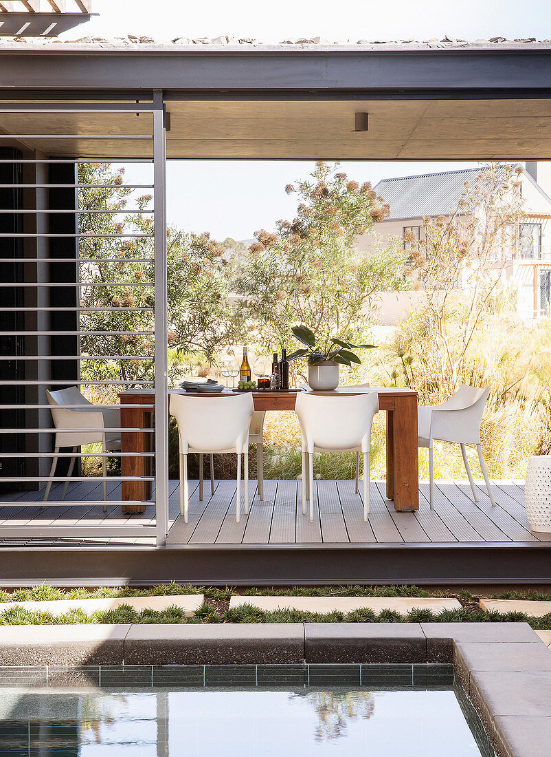 Dining table and white chairs on roofed terrace next to pool