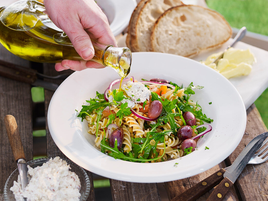 Olive oil being drizzled over a summer pasta salad with olives and rocket