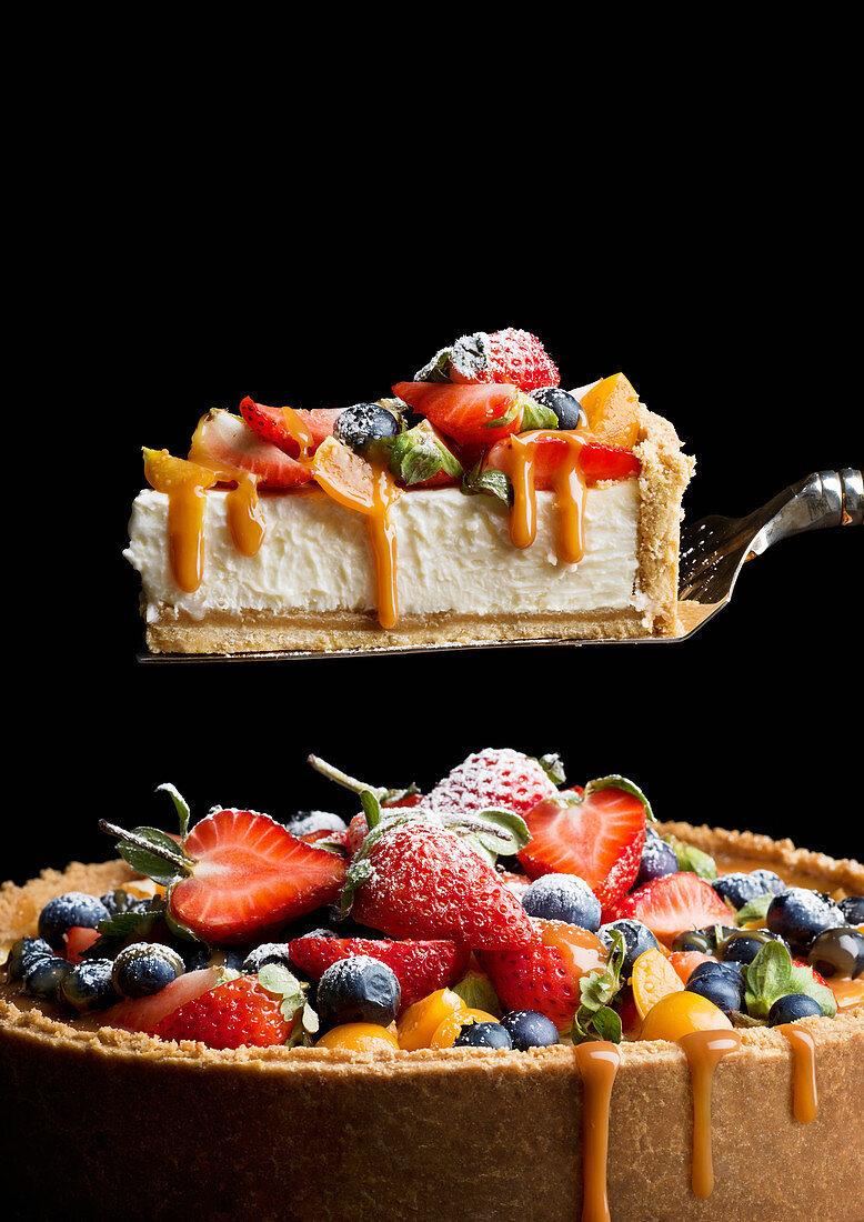 A cheesecake with caramel sauce and fresh fruit with a piece on a cake slice
