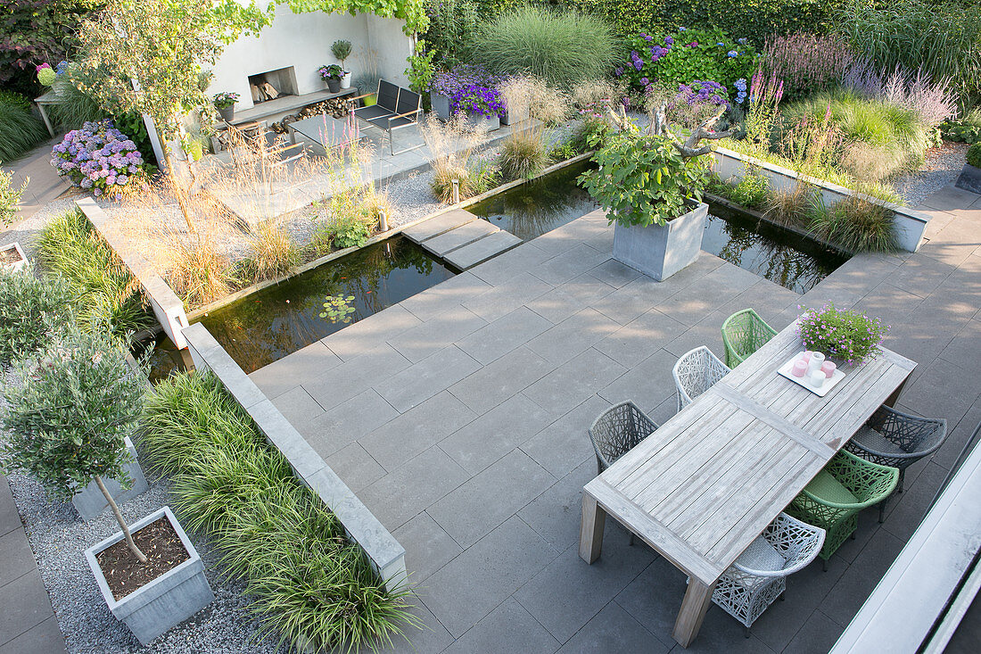 Wooden table and designer chairs on terrace with concrete flags surrounded by L-shaped pond