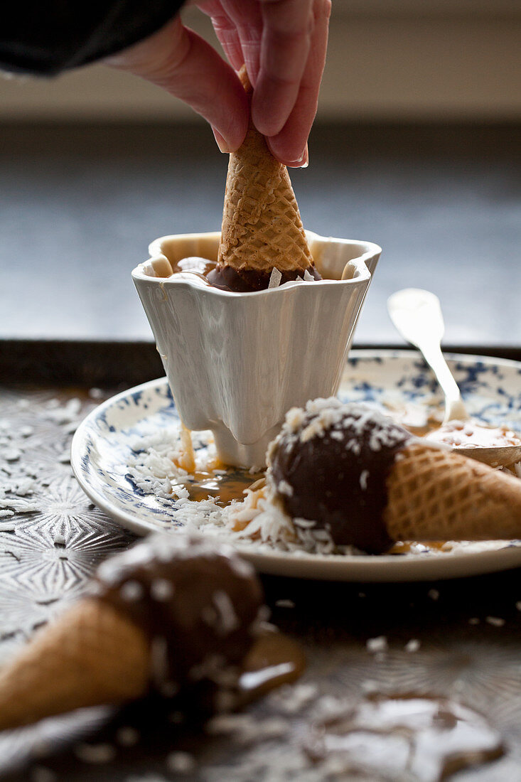 A chocolate covered ice cream cone being dipped into caramel sauce, with coconut and ice cream cones beside
