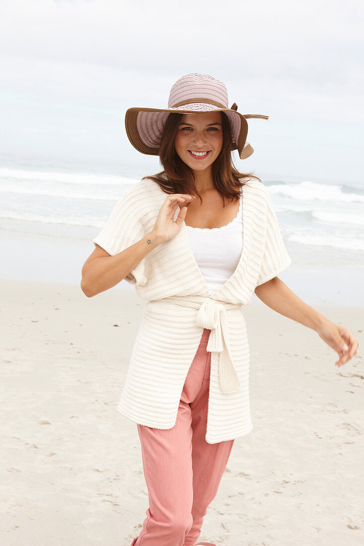 Brünette Frau mit Hut in Strickjacke mit kurzen Ärmeln und rosa Hose am Meer