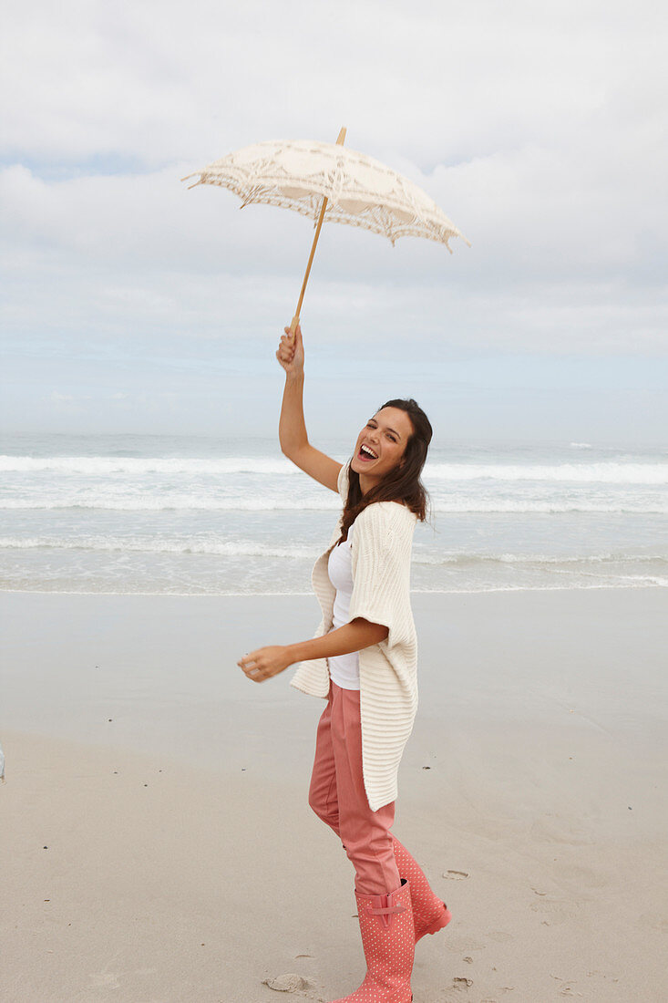 Brünette Frau mit Sonnenschirm in Strickjacke, rosa Hose und Gummistiefeln am Meer