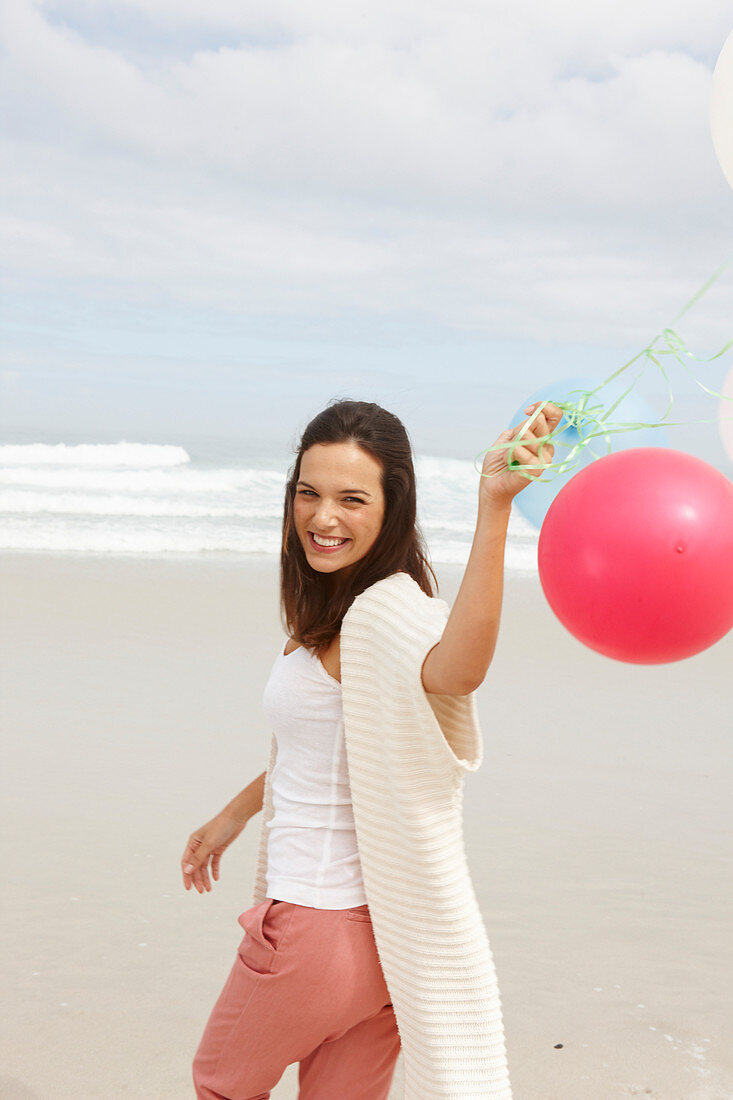 A brunette woman by the sea wearing a short-sleeved cardigan and holding balloons