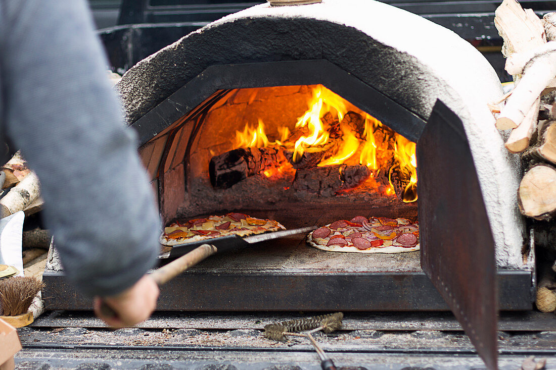https://media01.stockfood.com/largepreviews/MzkwMzkxODY2/12593286-Sliding-pizzas-into-a-small-portable-wood-fired-pizza-oven-with-live-flame-and-coals.jpg
