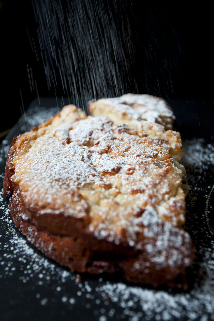 An apple cake on a black board, being dusted with powdered sugar