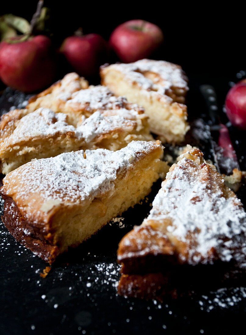 Apple cake slices, dusted with powdered sugar, on a black surface, with red apples