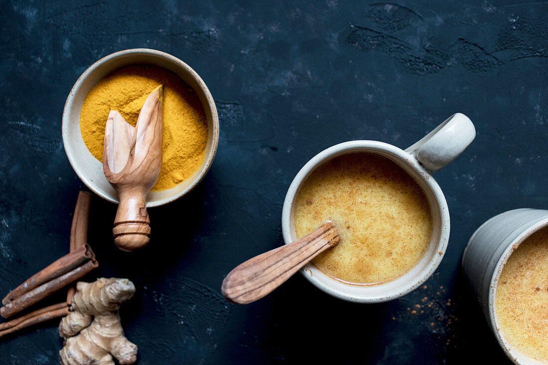 Bowl with turmeric and cinnamon with ginger lying near cups of fresh latte on dark tabletop