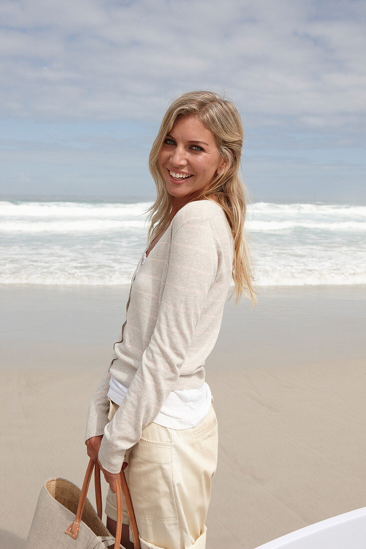 Blonde Frau mit Tasche in hellem Cardigan und Shorts am Strand