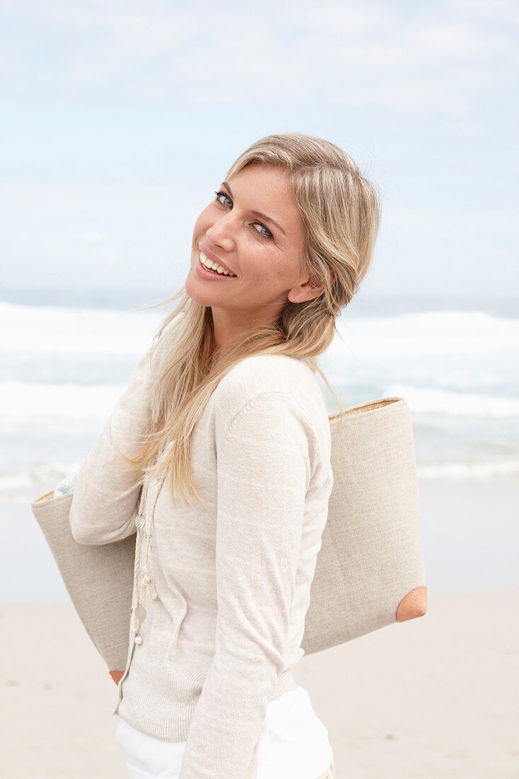 A blond woman on a beach wearing a light cardigan and shorts and holding a bag