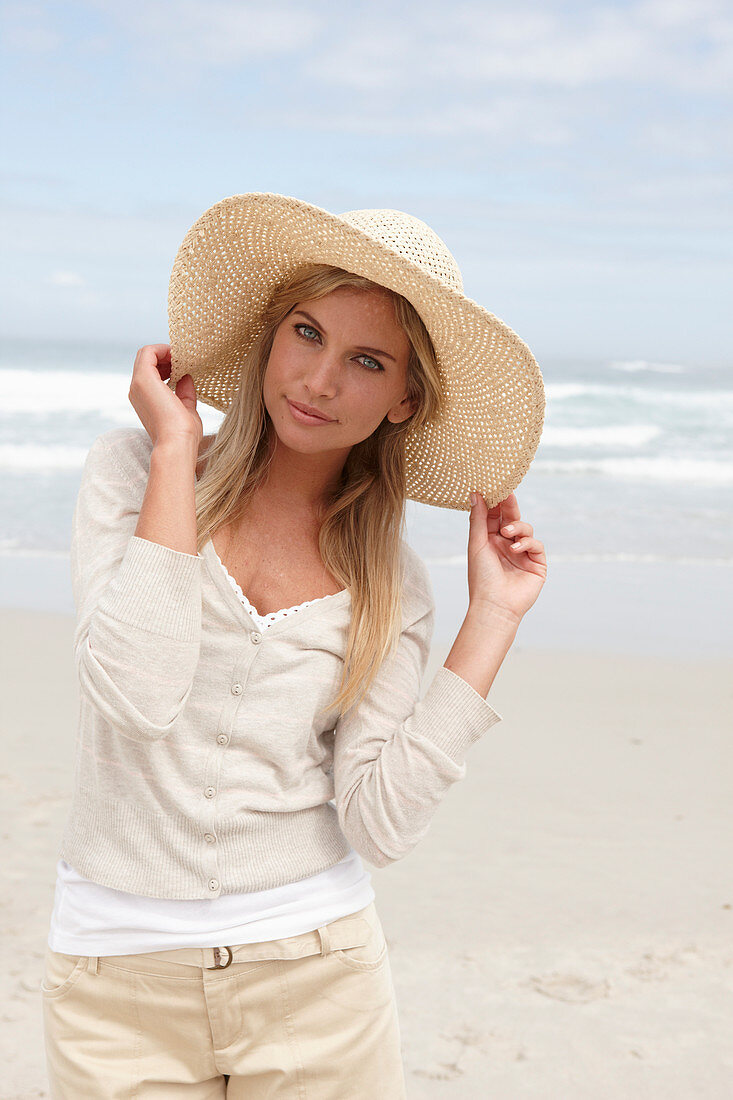 A blond woman on a beach wearing a light cardigan, shorts and a summer hat