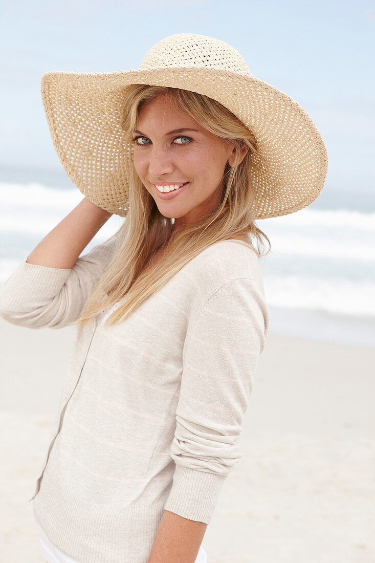 Blonde Frau mit Sommerhut in hellem Cardigan am Strand