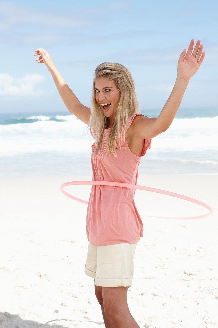 Junge Frau mit Hula-Hoop-Reifen im rosa Top und Shorts am Strand