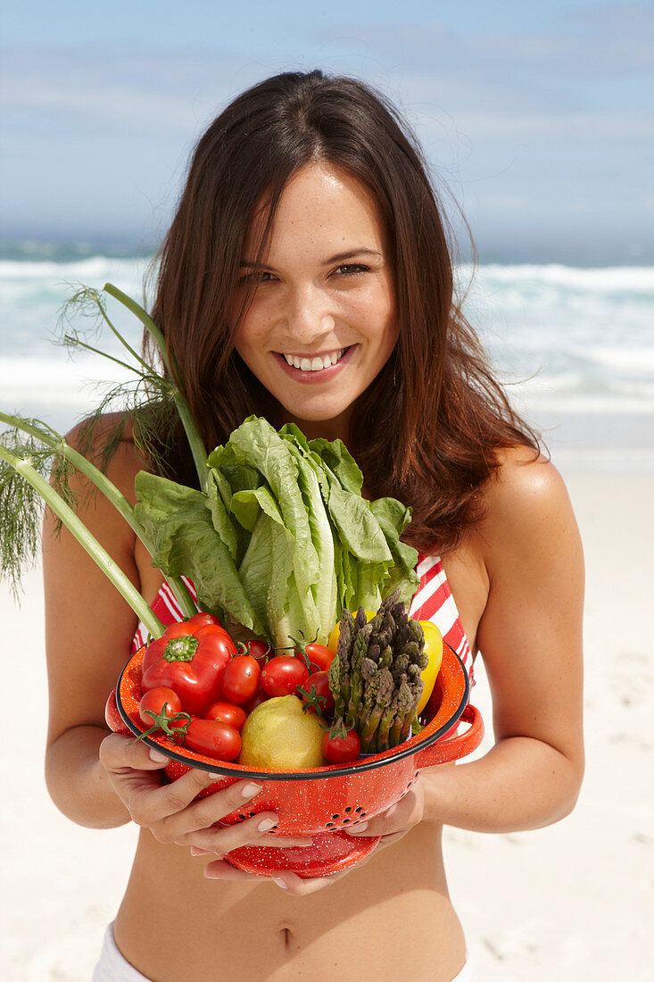 A young brunette woman on a beach wearing a bikini top and holding a bowl of vegetables