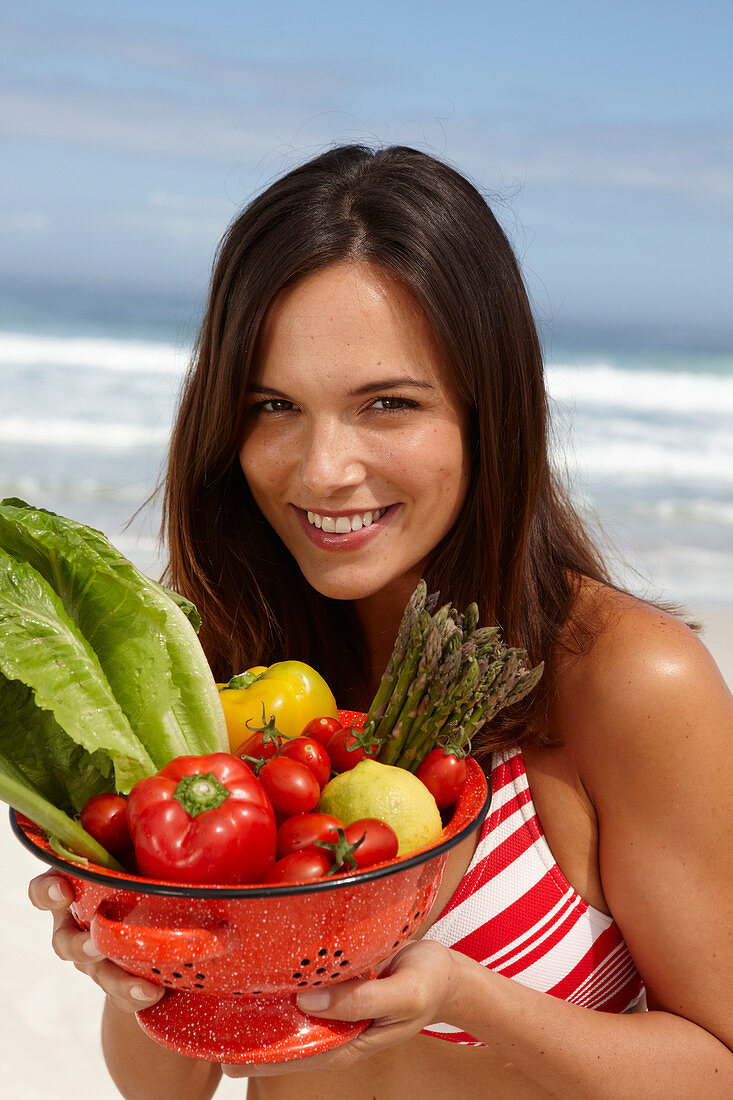 A young brunette woman on a beach wearing a bikini top and holding a bowl of vegetables