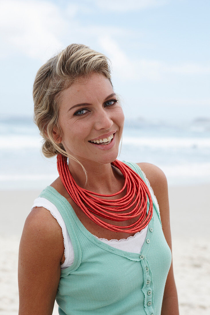 A blonde woman on a beach wearing a turquoise top and a red necklace