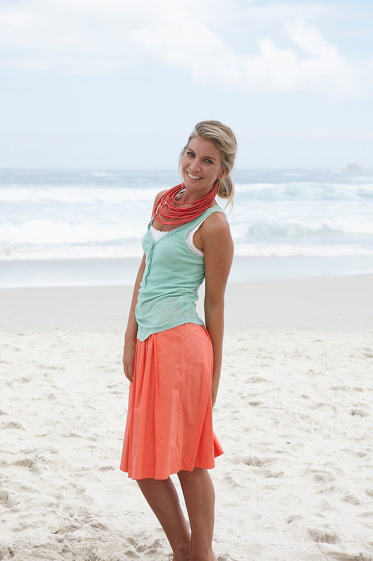 A blonde woman on a beach wearing a turquoise top, a red necklace and a salmon-pink skirt