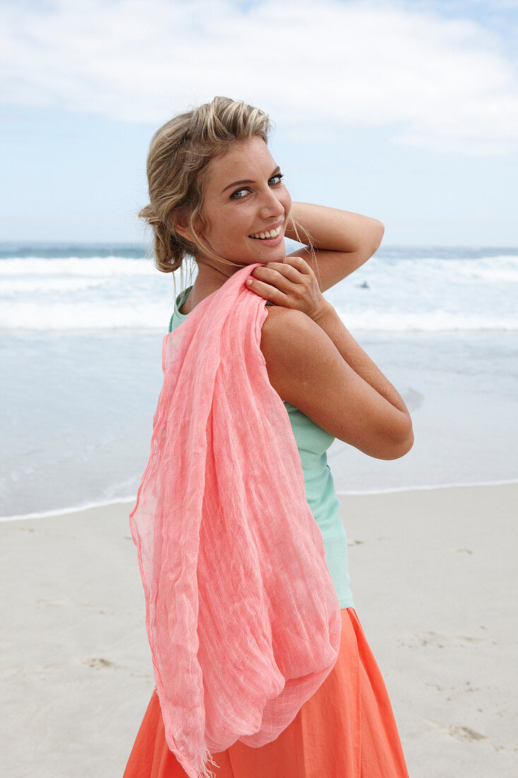 A blonde woman on a beach with a scarf wearing a turquoise top and a salmon-pink skirt