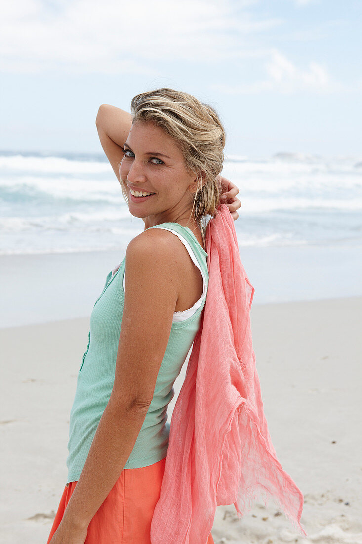 A blonde woman on a beach with a scarf wearing a turquoise top and a salmon-pink skirt