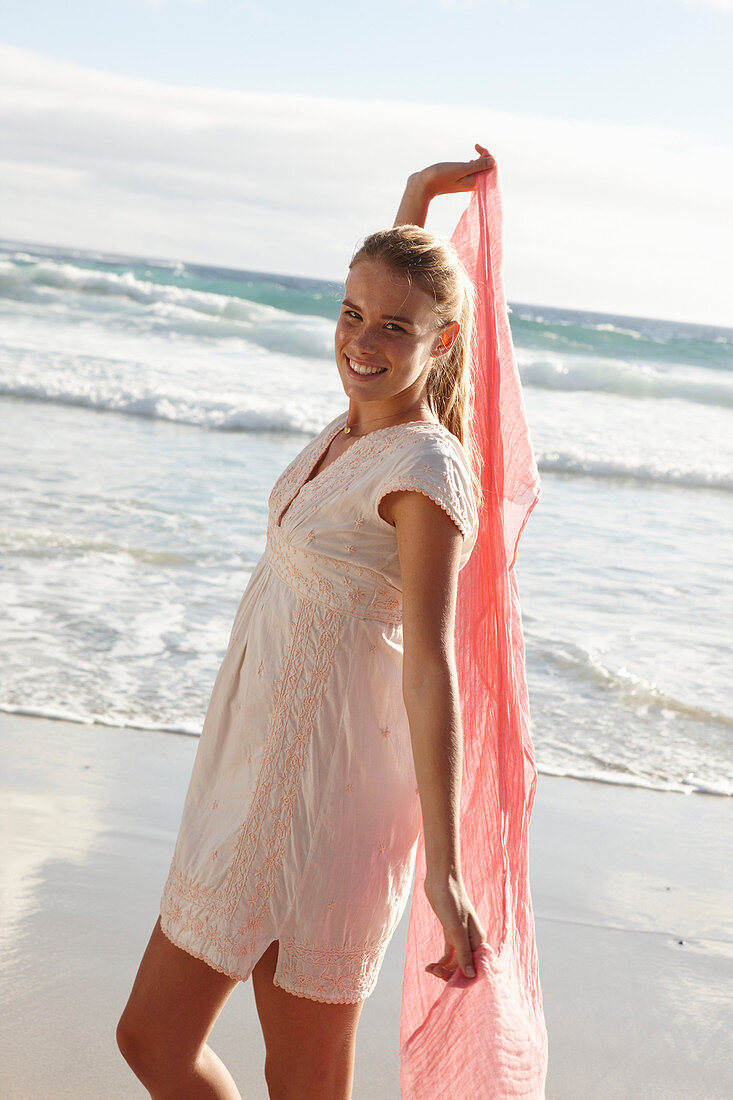 A blonde woman by the sea with a salmon-pink scarf and wearing a white dress