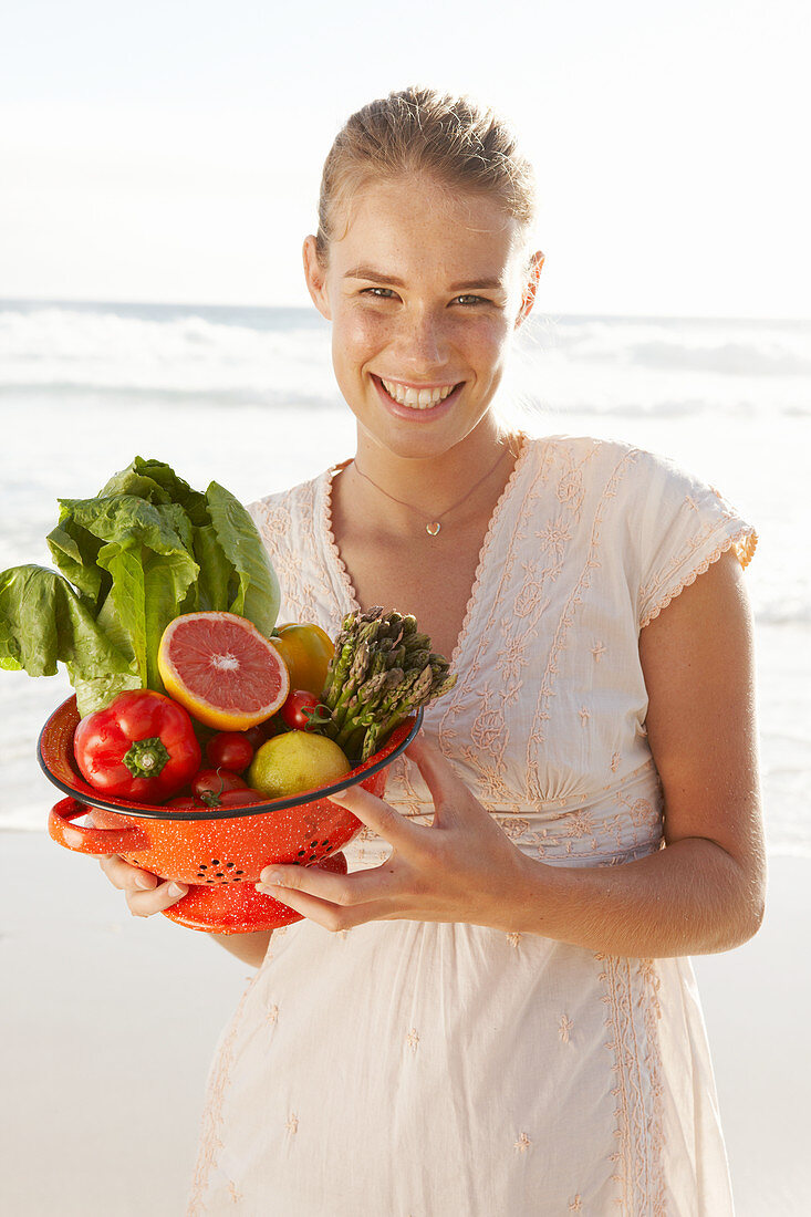 A blonde woman by the sea with a bowl of fruit and vegetables wearing a white dress