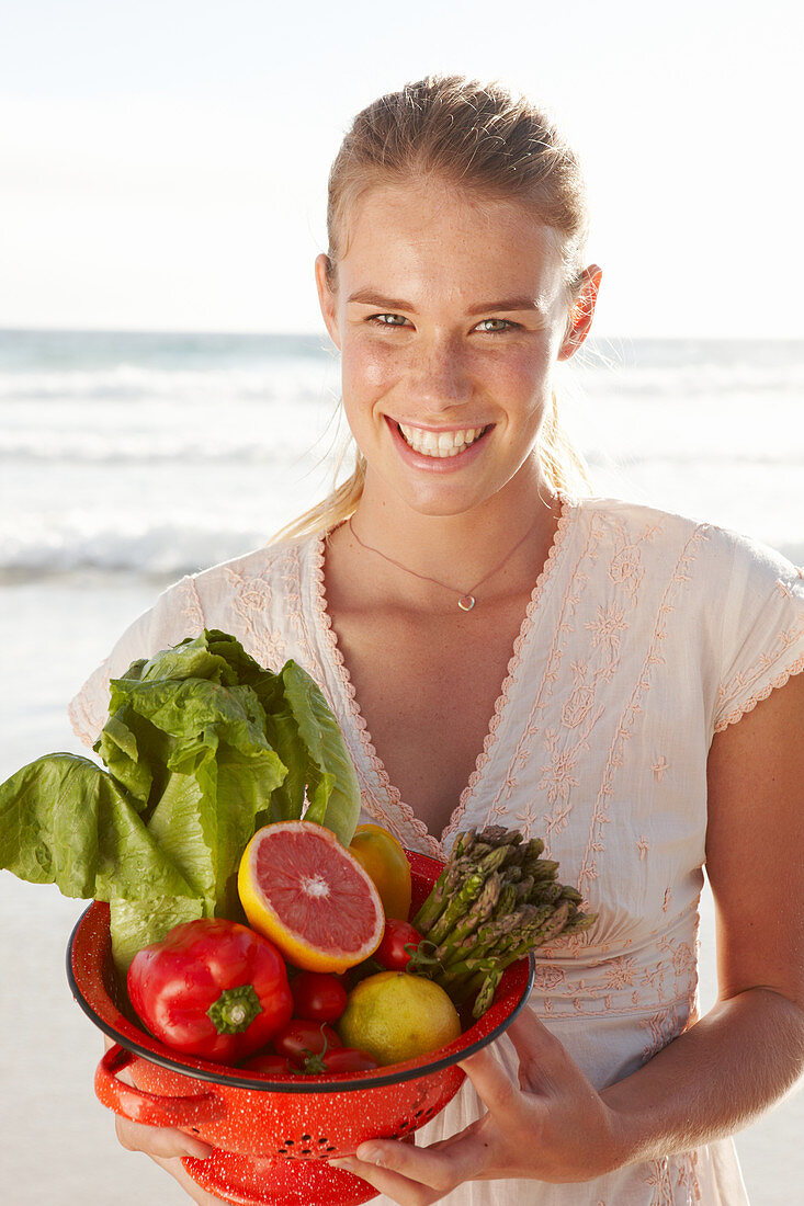 A blonde woman by the sea with a bowl of fruit and vegetables wearing a white dress