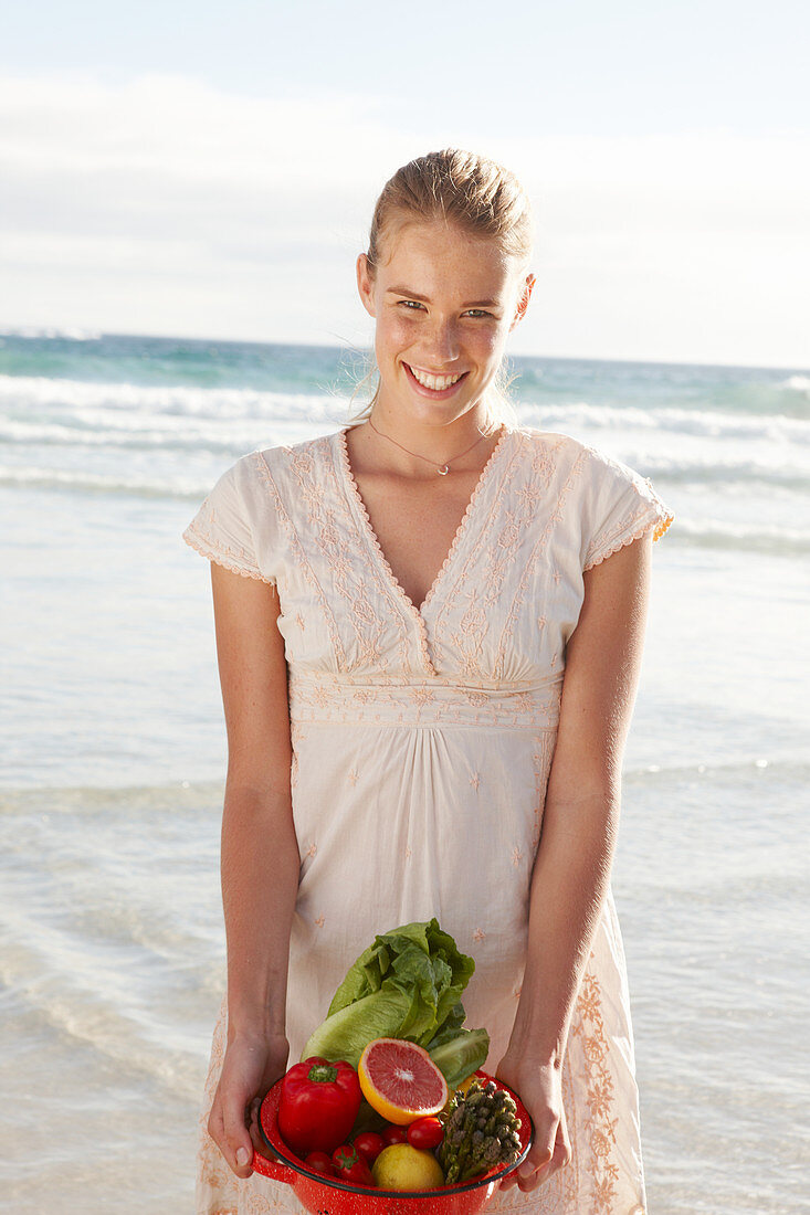 A blonde woman by the sea with a bowl of fruit and vegetables wearing a white dress