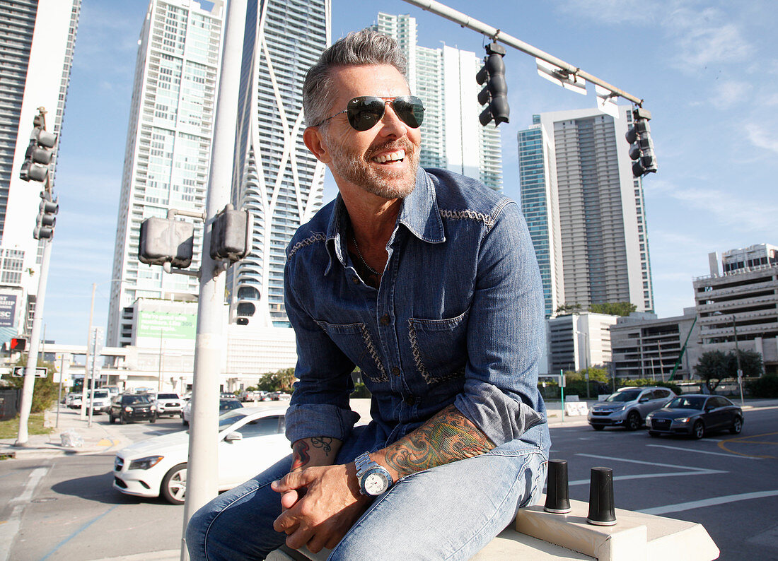 A man with a bread wearing a denim shirt and jeans sitting against a skyline backdrop
