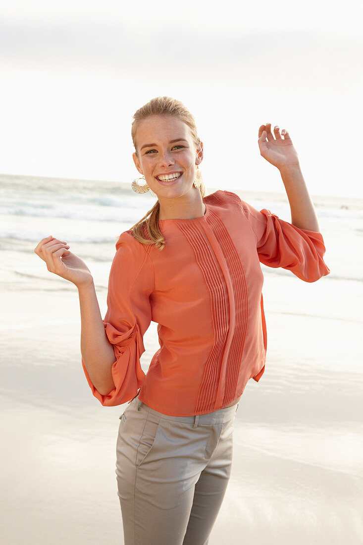 A young blonde woman wearing an orange blouse and light trousers by the sea