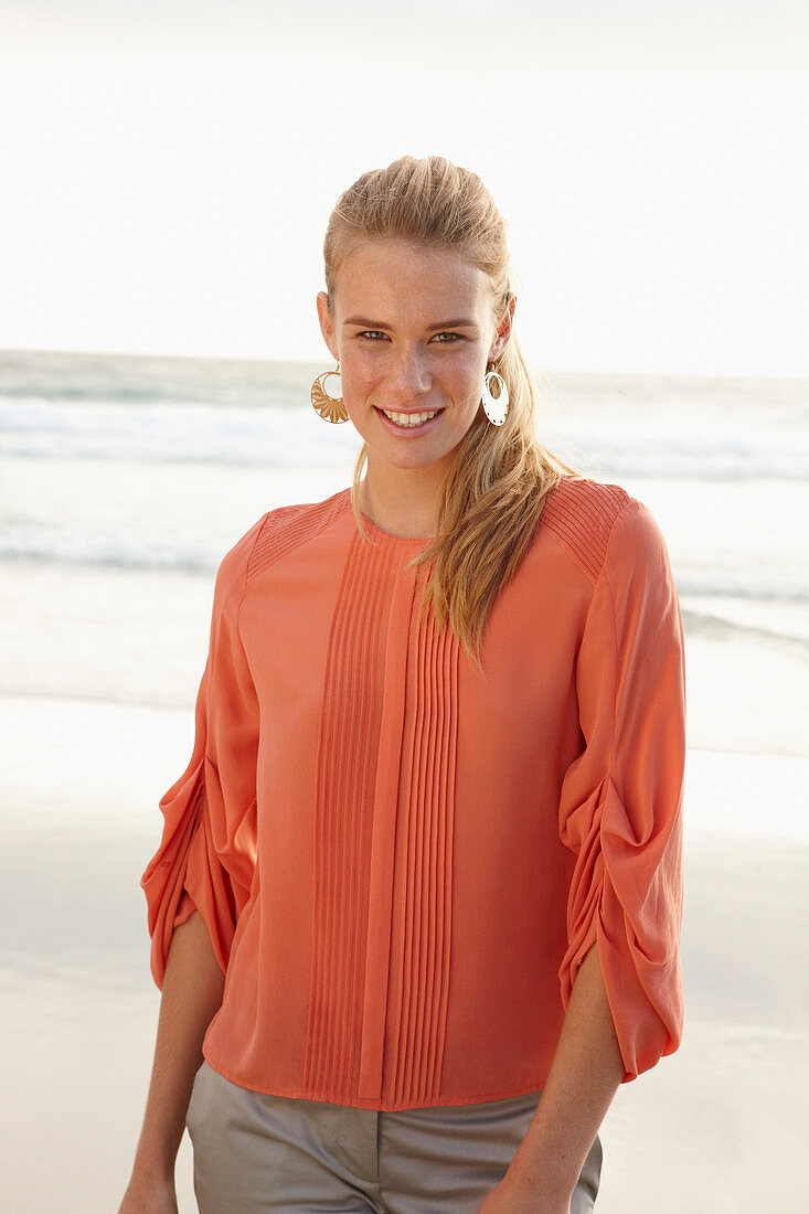 A young blonde woman wearing an orange blouse and light trousers by the sea
