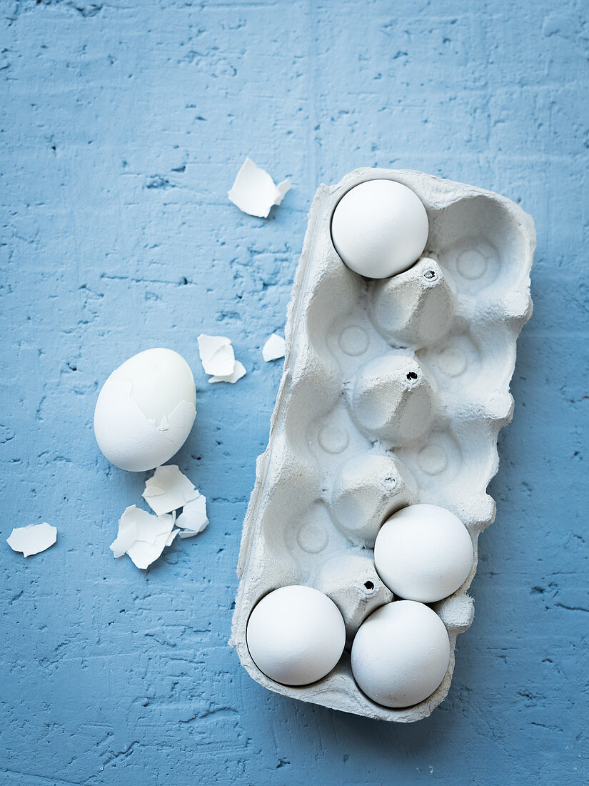 weiße Eier in Eierkarton, daneben gekochtes teilweise gepelltes Ei