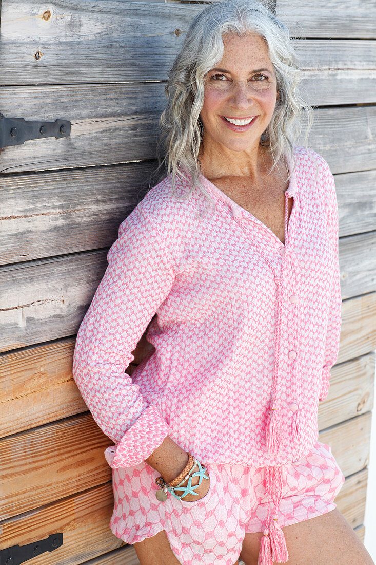 A mature woman wearing a pink blouse and pink shorts standing against a wooden wall