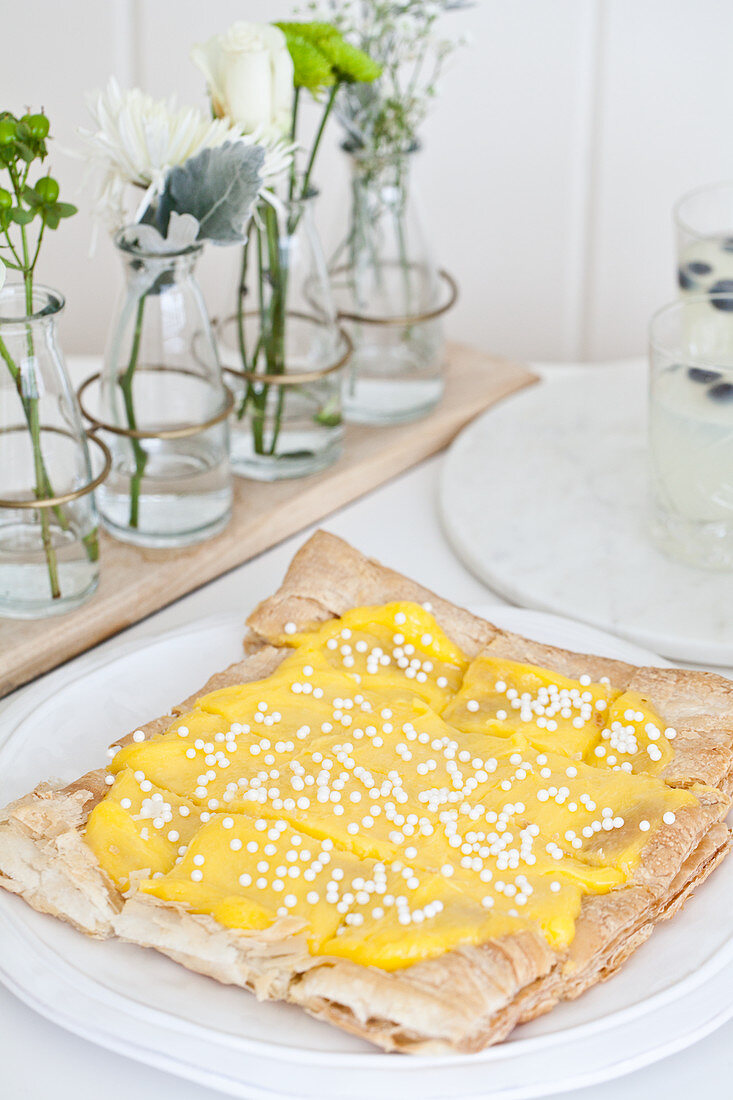 Easter lemon curd puff pastry tart topped with sugar pearls on a white plate with flowers and lemonade with blueberries