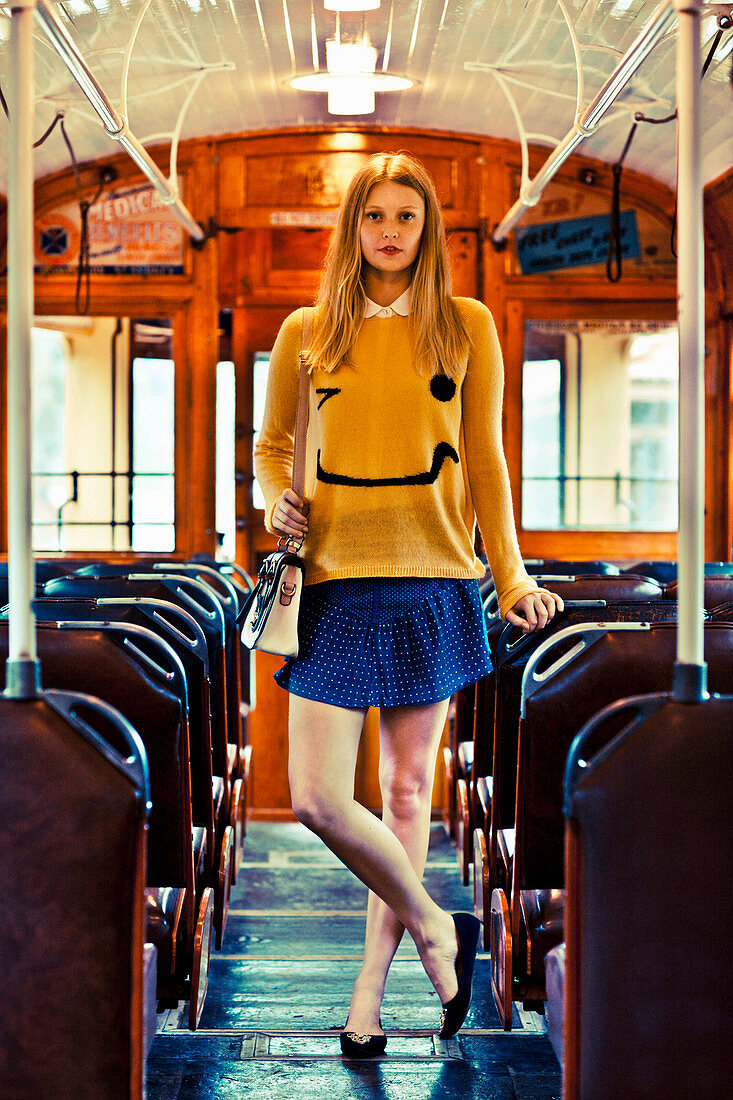 A young blonde woman wearing a smiley jumper and a blue skirt standing on a bus